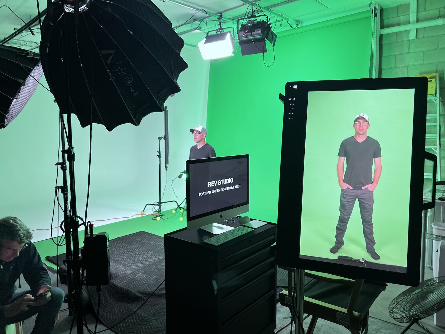 REV Studios green screen stage with lighting setup, a subject standing on screen, and an iMac displaying the studio name.